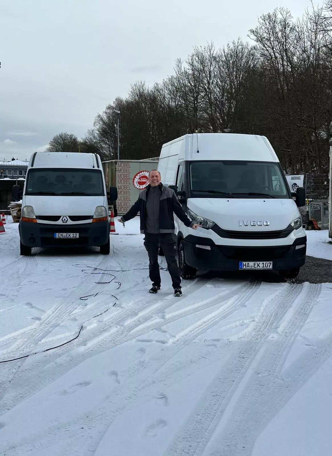 Ein Mitarbeiter steht lächelnd zwischen zwei weißen Transportern im Schnee.